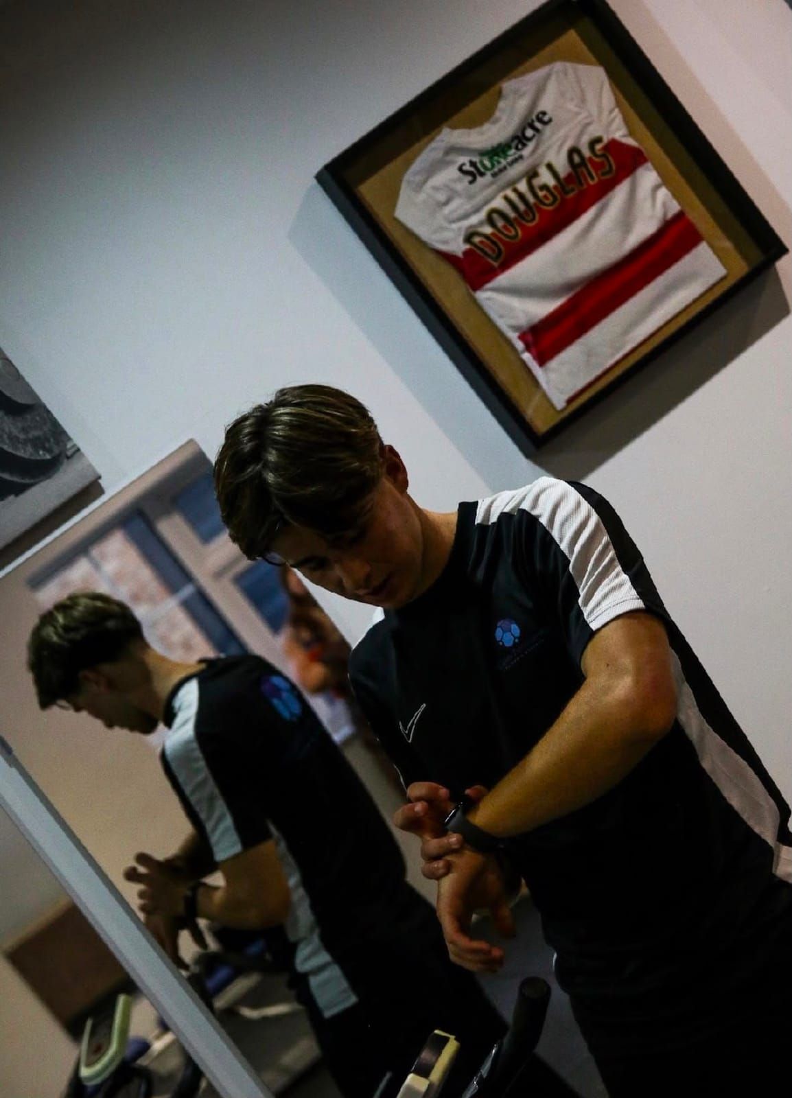 A coach in HDperformance kit checks his watch in the gym, beneath a framed football shirt