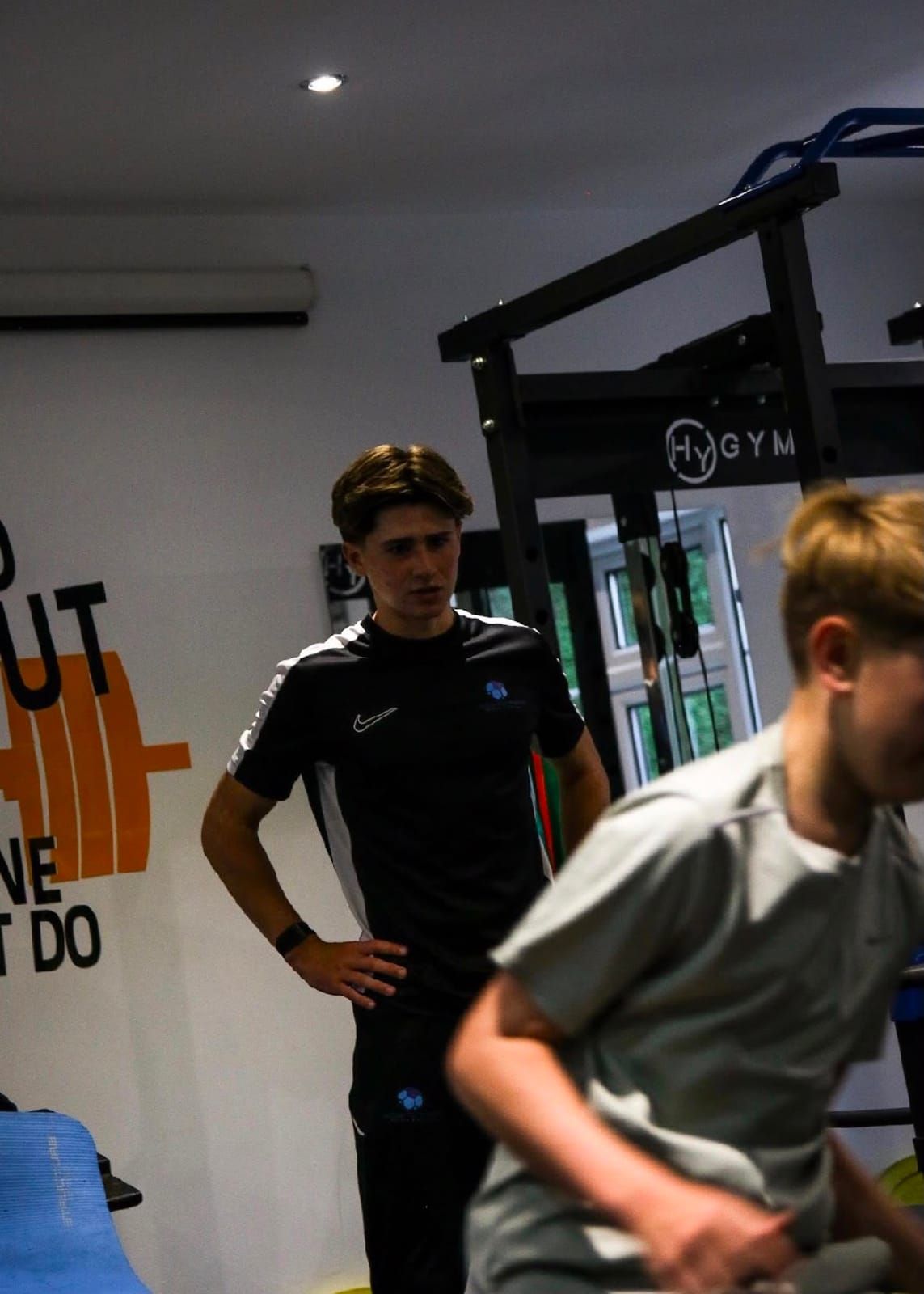 A coach watches closely as a young athlete moves through an exercise in front of a power rack