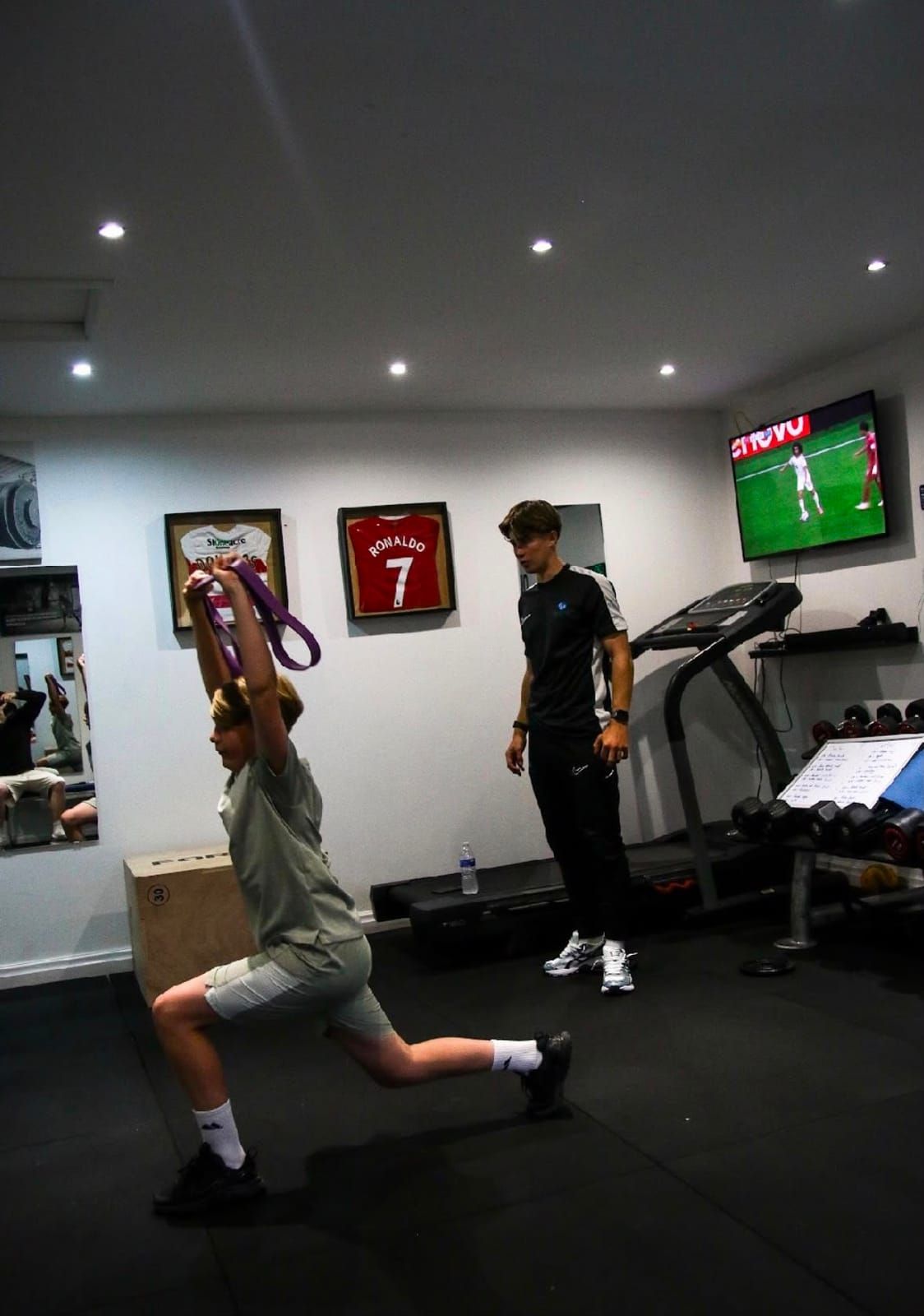 A young athlete performs a lunge with a resistance band overhead while a coach watches, in a gym with mats, dumbbells and framed football shirts