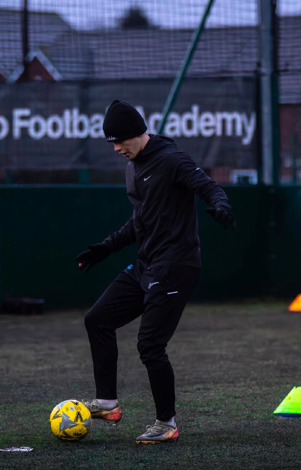 A coach in a black hat and gloves works the ball with the sole of his boot on a 4G pitch, with cones nearby