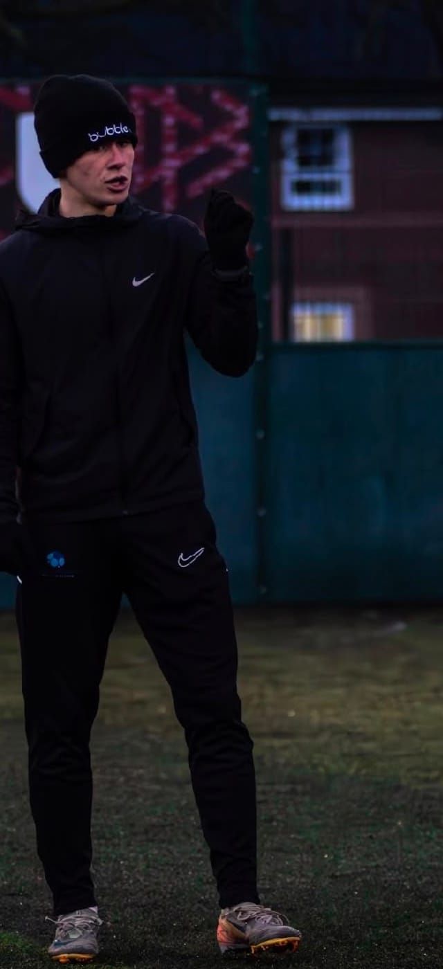 A coach in a black hat and training kit gives instructions on a 4G pitch at dusk