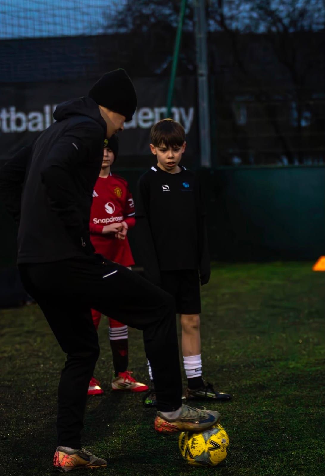 A coach demonstrates a move with his foot on the ball while two young players watch closely on a 4G pitch