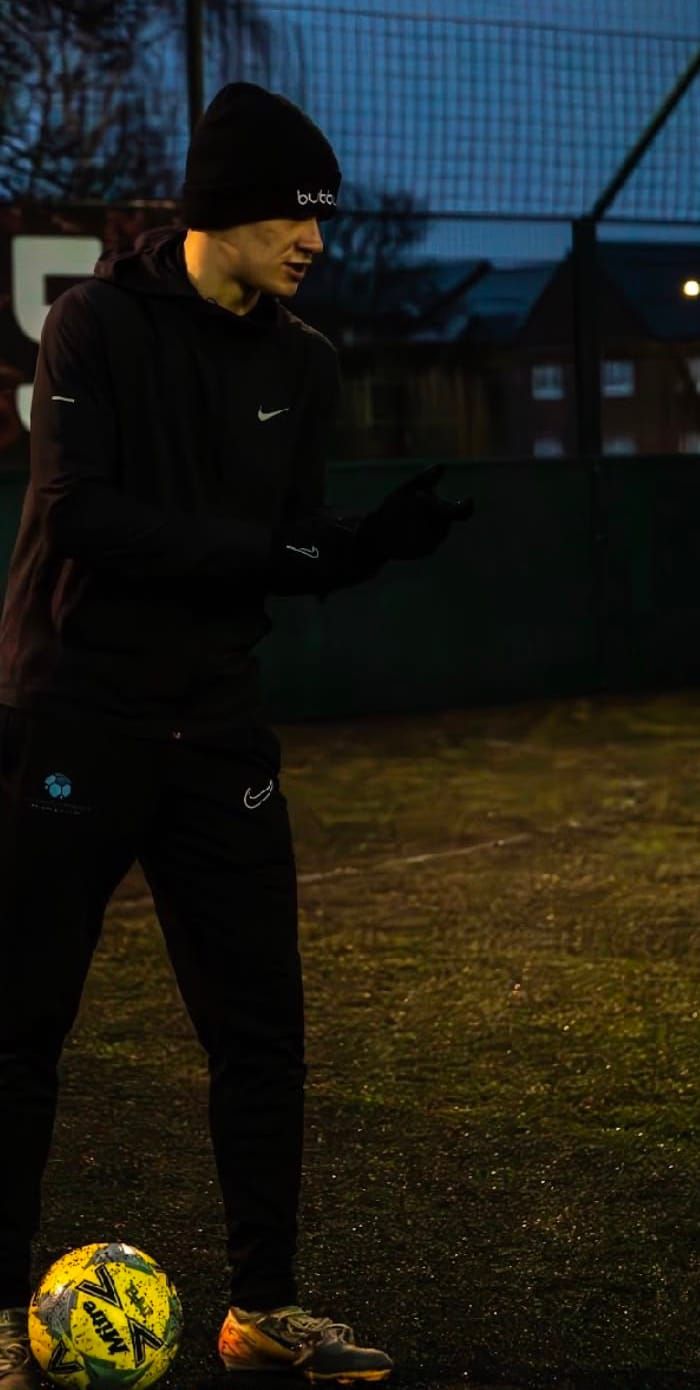 A coach in a black hat stands beside a yellow football on a 4G pitch at dusk, explaining the next drill