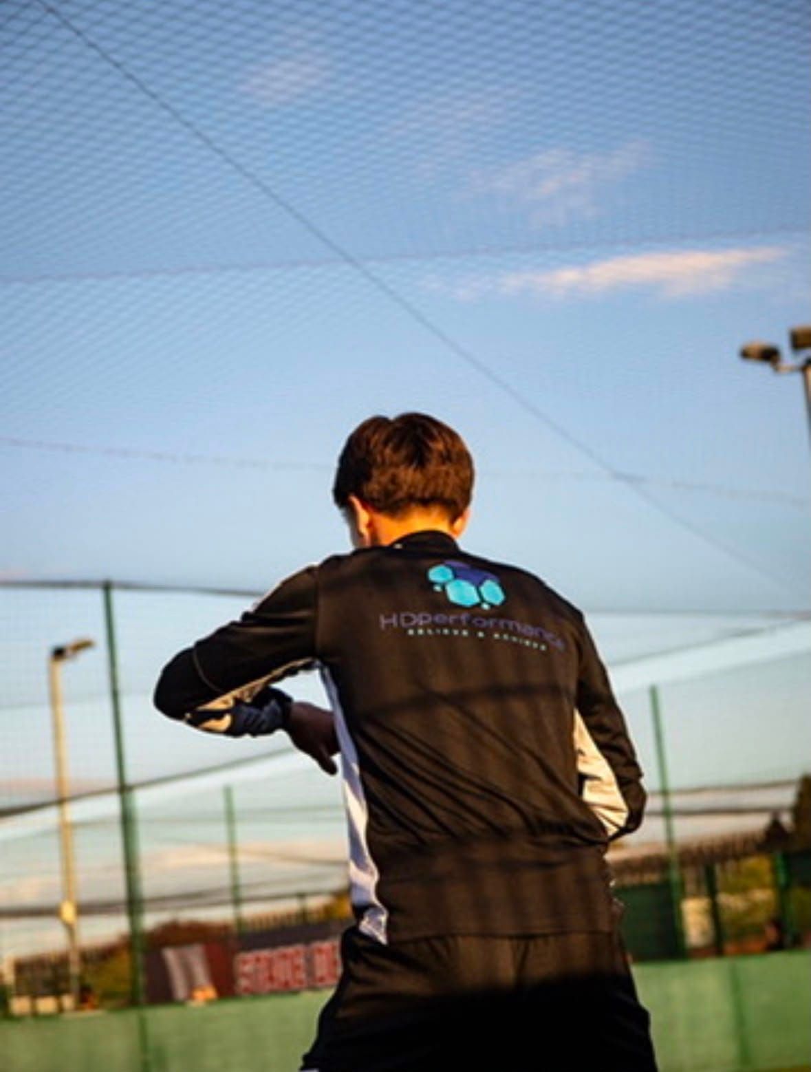A player seen from behind wearing a black HDperformance training top, on a netted outdoor pitch under a blue sky