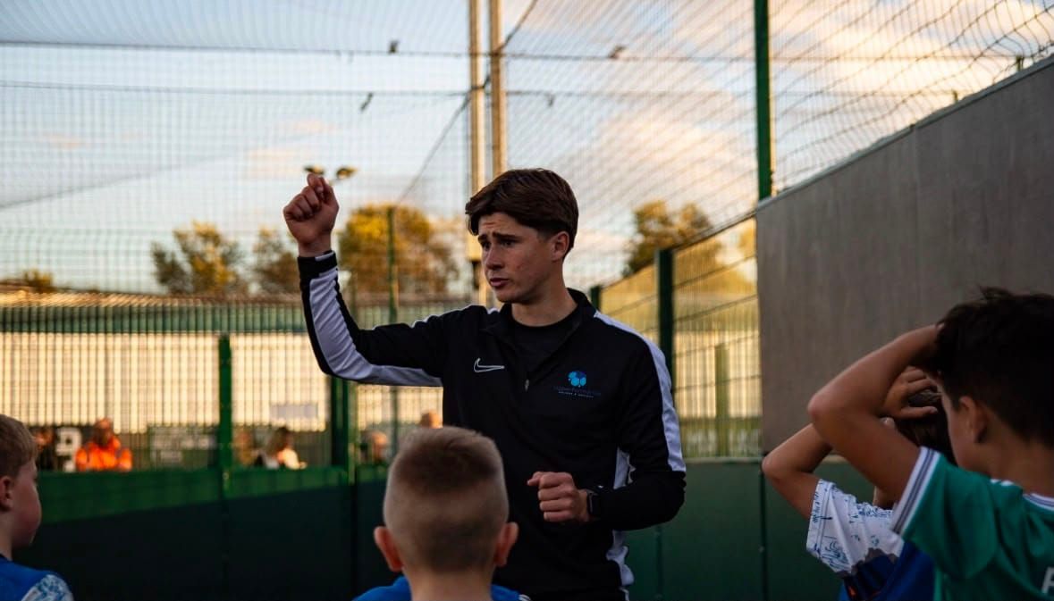 A coach in an HDperformance training top speaks to a group of young players gathered around him on a caged pitch