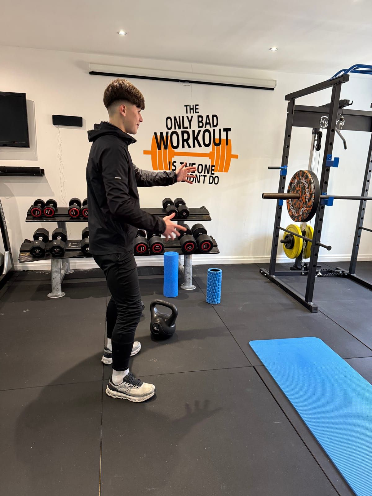 A coach in the youth performance gym, with dumbbells, a power rack, foam rollers and a blue exercise mat