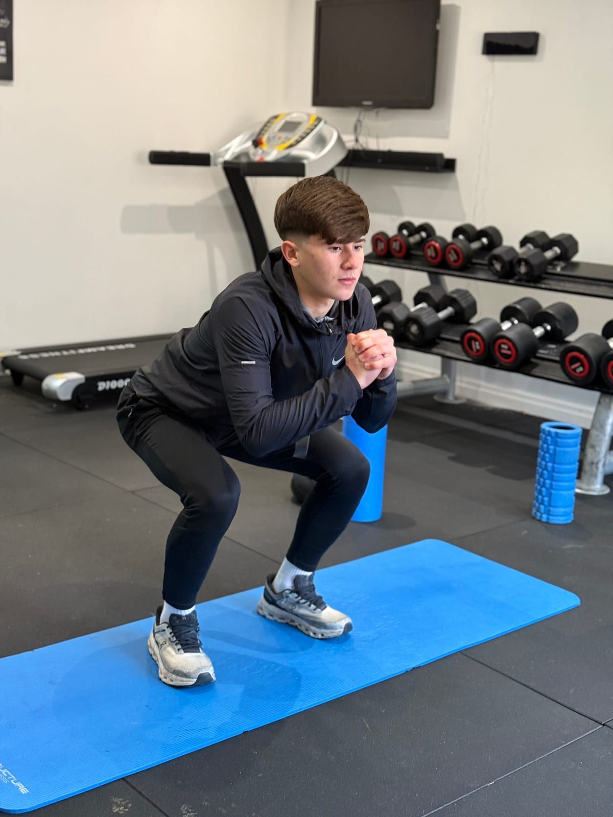 A coach demonstrates a bodyweight squat on a blue mat in the youth performance gym