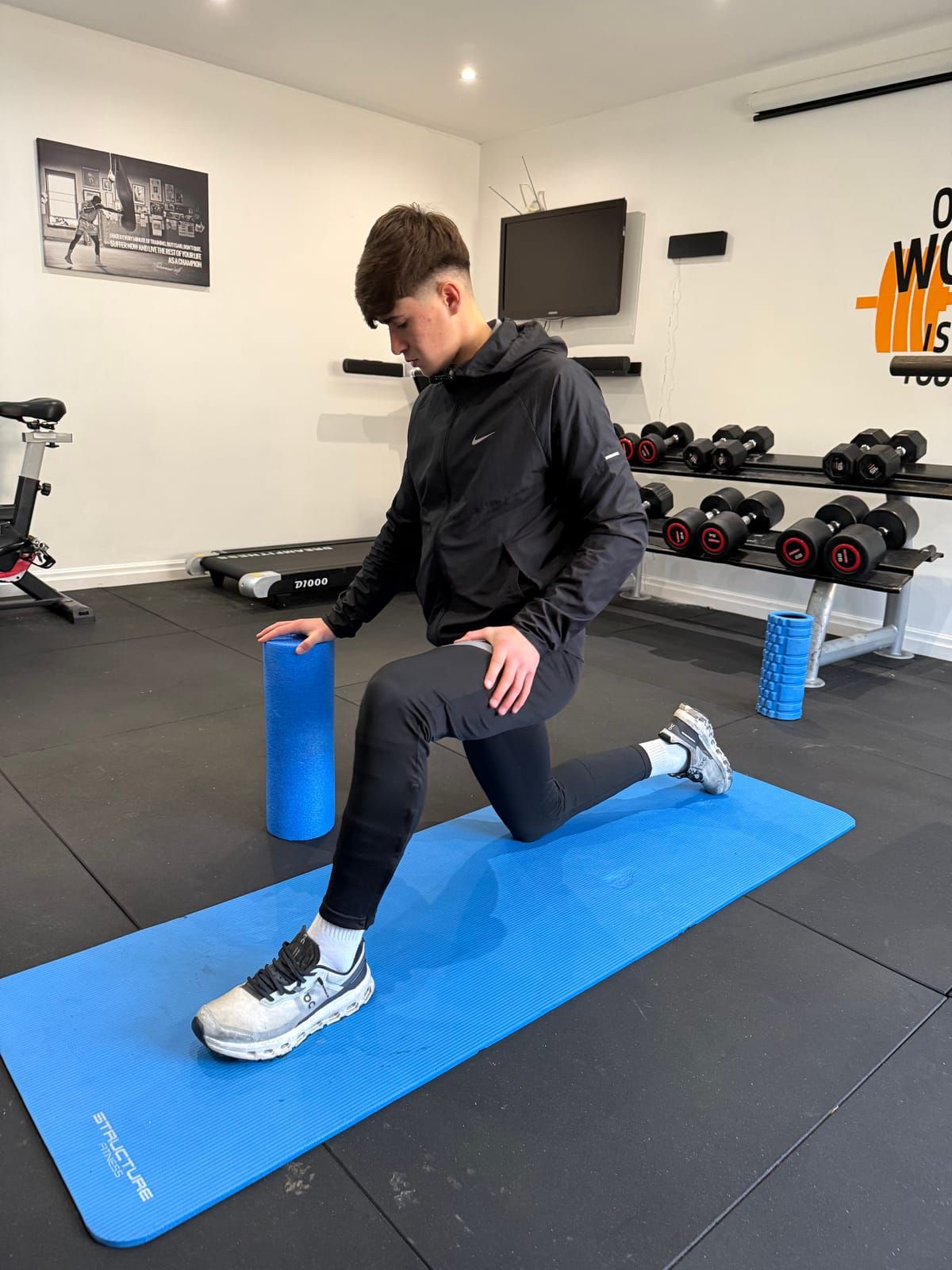 A coach demonstrates a kneeling lunge with a foam roller on a blue mat in the gym