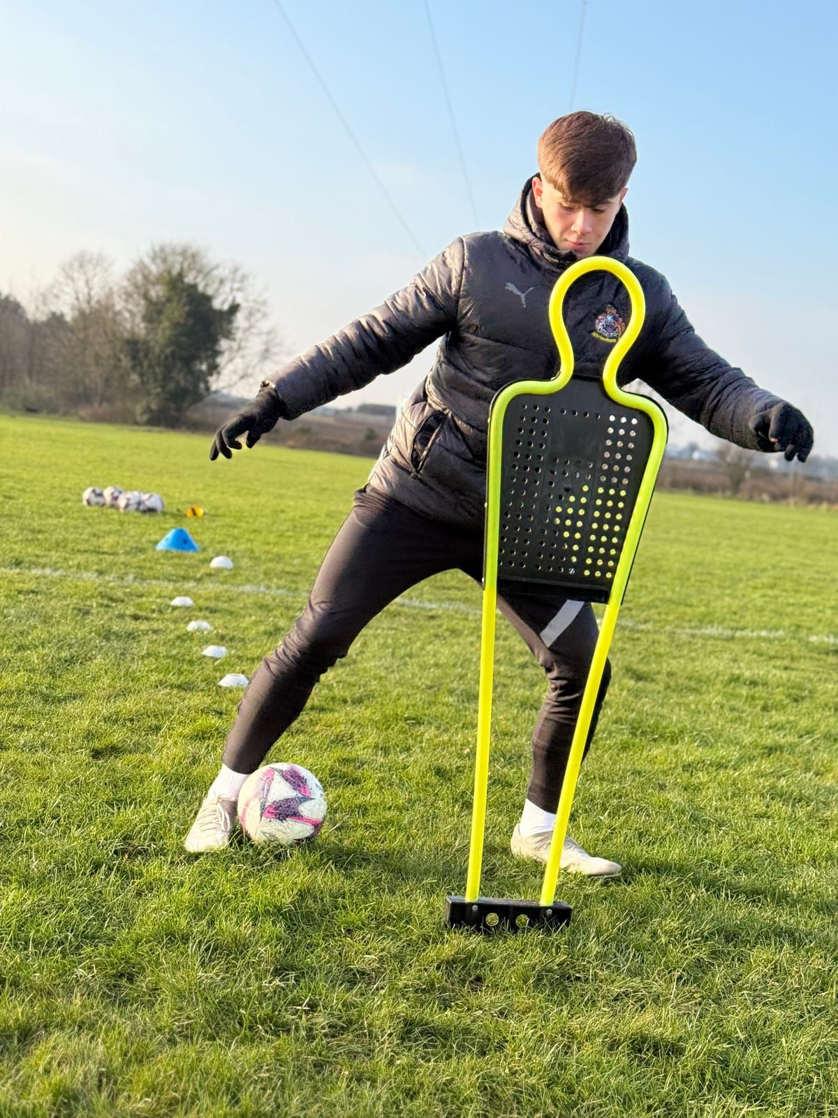 A coach in a black jacket dribbles a football past a yellow training mannequin on a grass pitch