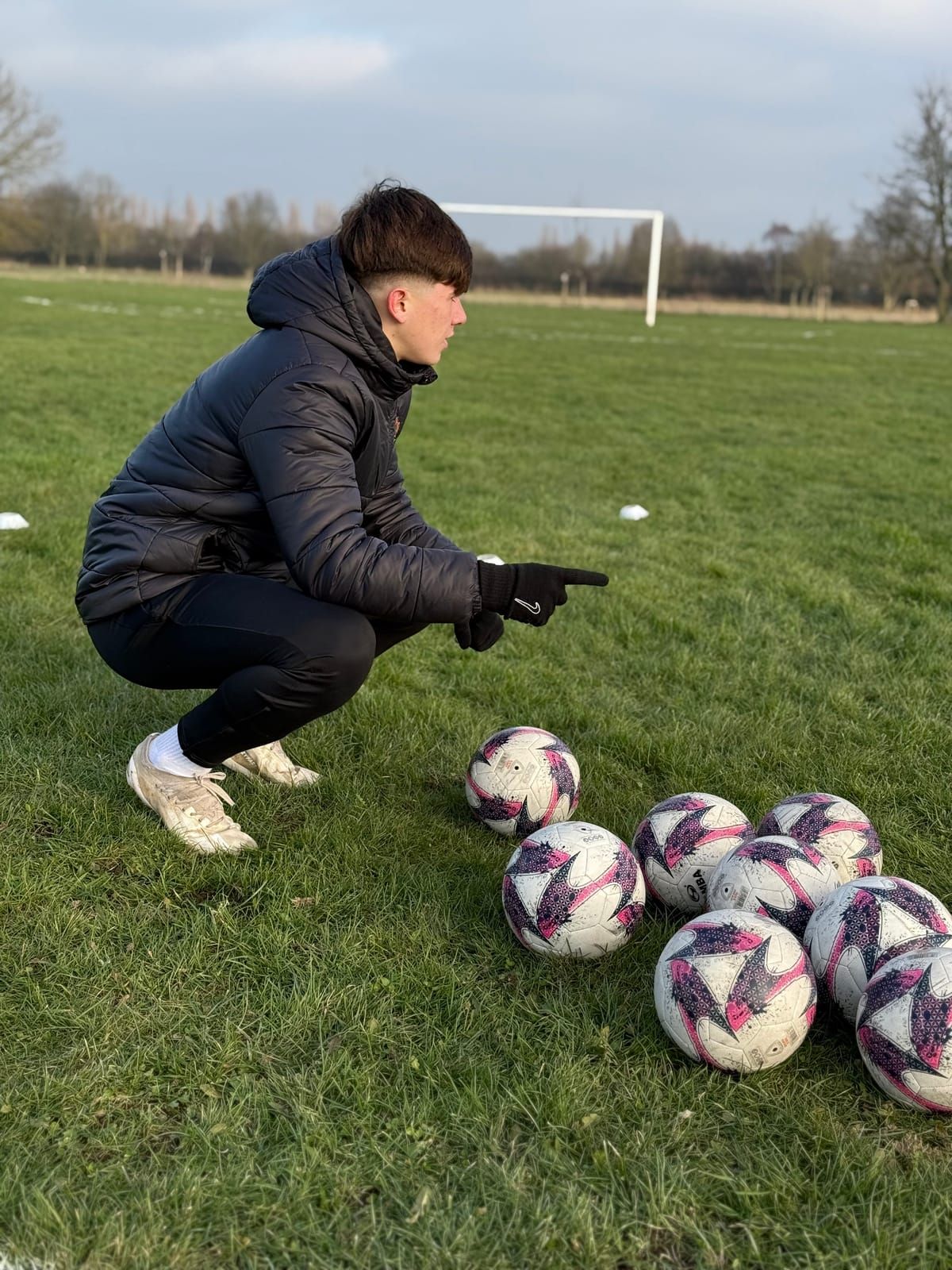 A coach crouches beside a pile of footballs on a grass pitch, pointing as he explains a drill