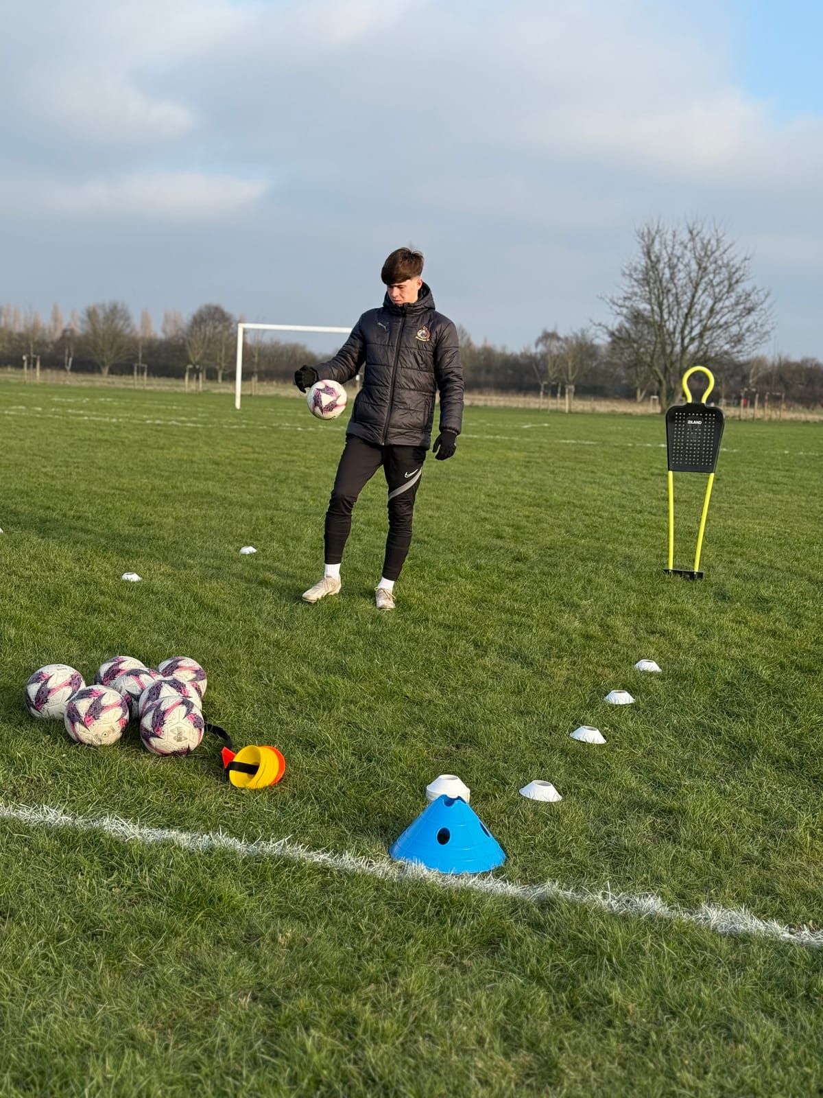 A coach with a ball stands on a wide grass pitch set out with cones, footballs and a mannequin, a goal behind him
