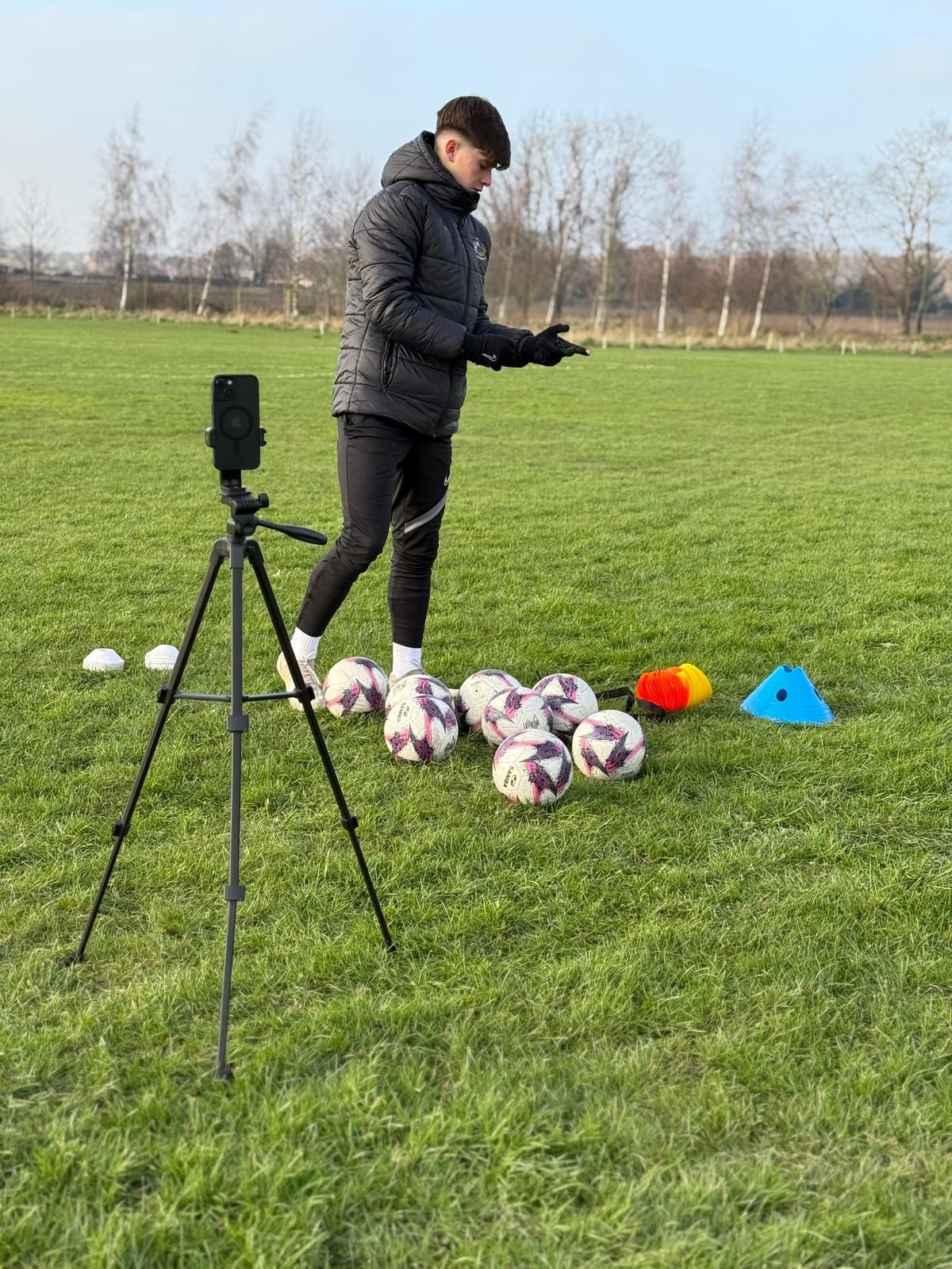 A coach sets up a drill beside a phone on a tripod, with footballs and cones on the grass