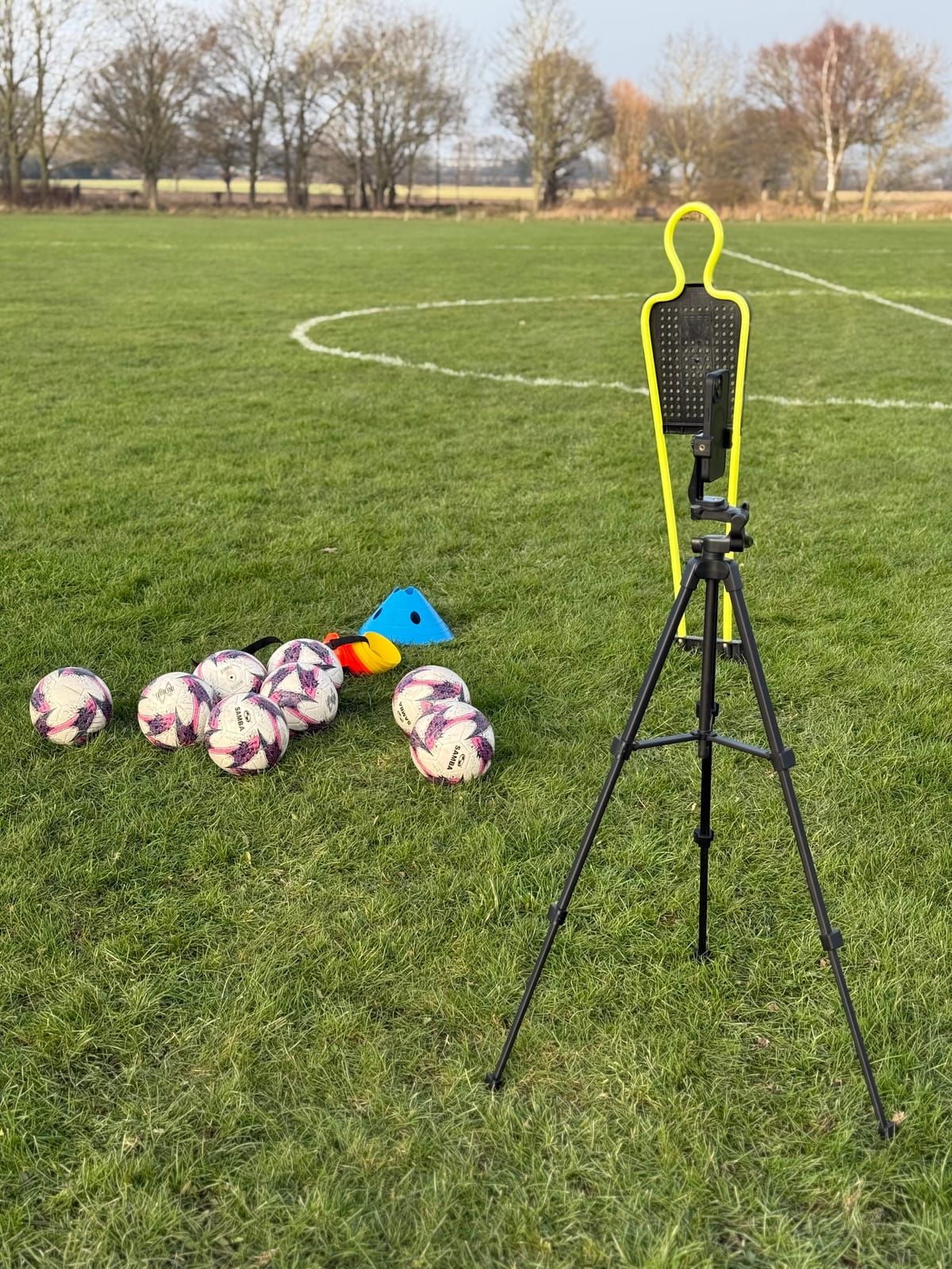 Footballs, cones, a camera tripod and a yellow mannequin laid out on a grass pitch before a session
